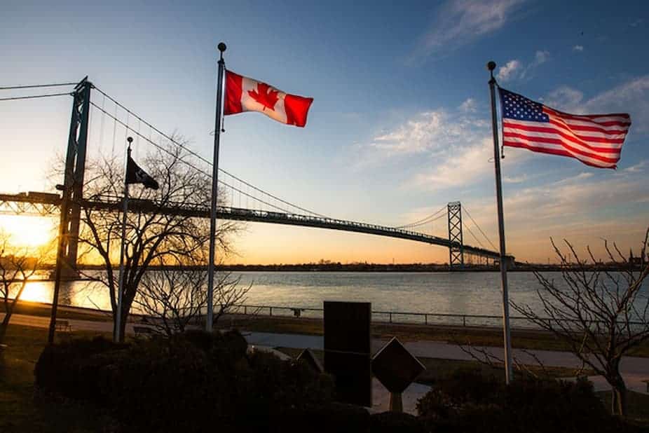 U.S. and Canadian flags flying beside a bridge over a river at sunset.