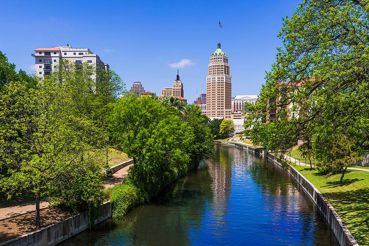 Tree-lined riverwalk with a calm canal and downtown buildings under a clear blue sky