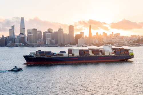 Container ship sailing across the bay with a city skyline at sunset in the background.